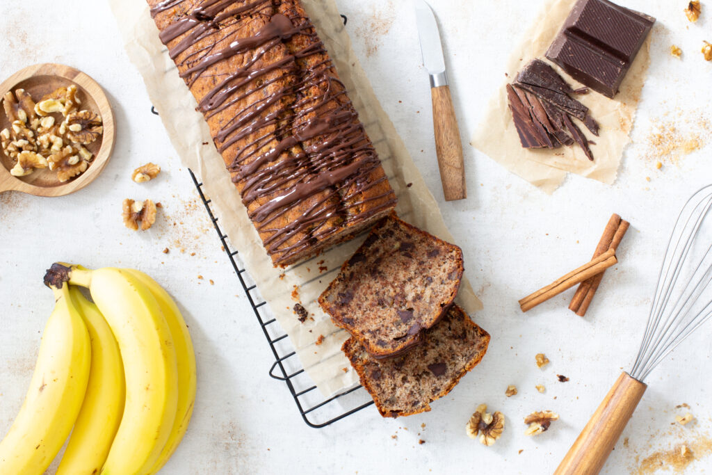 Bananenkuchen aka Bananenbrot als Kastenkuchen mit aufgeschnittenen Stücken von oben fotografiert. Nebendran liegen ein Schneebesen, ein Messer, Bananen, Walnüsse, Zimtstangen und ein großes Stück Schokolade