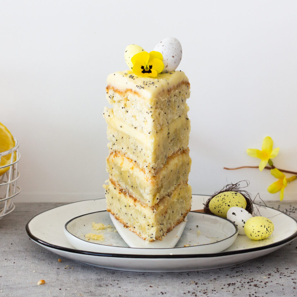 Ein Stück zitronig-frische Ostertorte mit Mohn und farbenfroher Frühlingsdeko on top