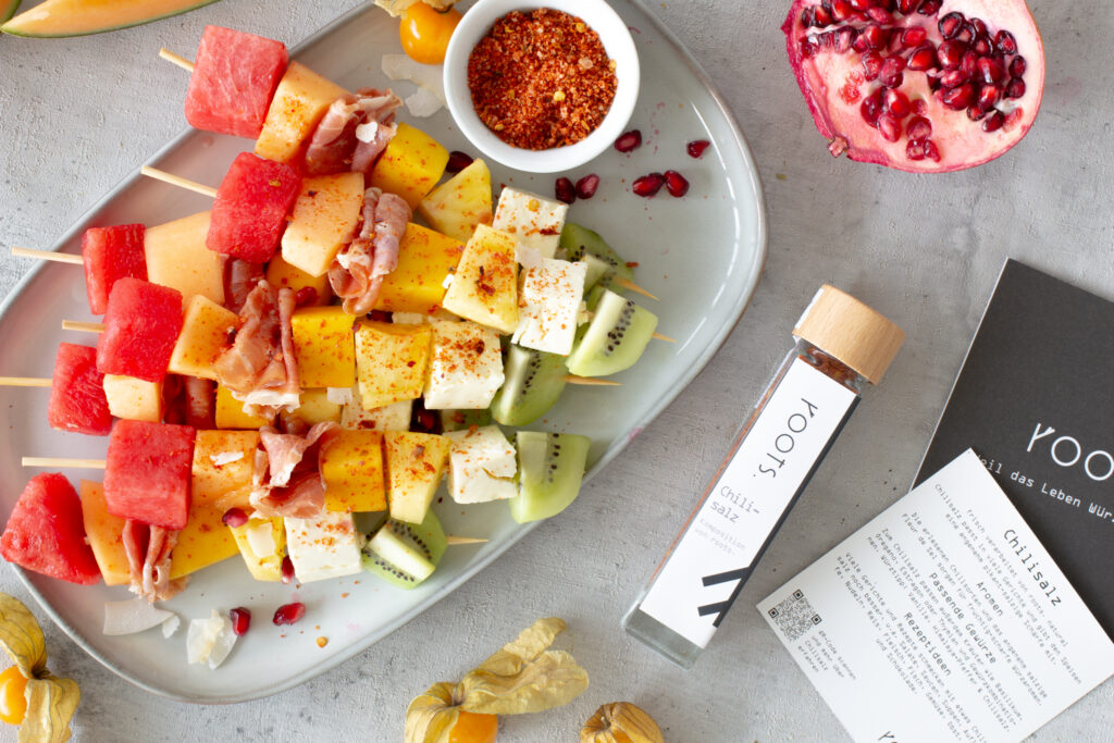 Mehrere exotische Obstspieße aus Melone, Mango, Ananas, Kiwi, Feta und Parmaschinken auf einem Teller. Nebendran liegt das Chilisalz von Roots mit dem die Obstspieße gewürzt sind