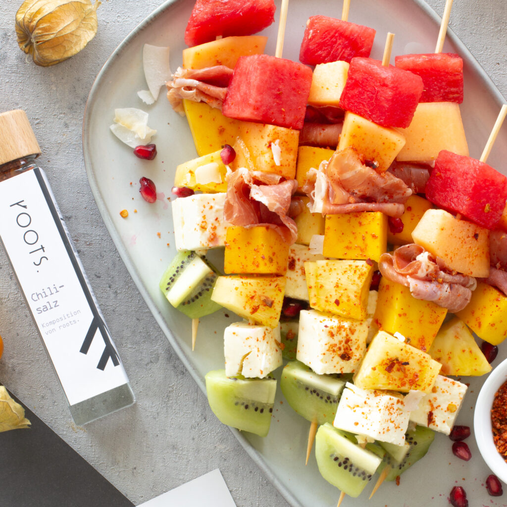 Mehrere exotische Obstspieße aus Melone, Mango, Ananas, Kiwi, Feta und Parmaschinken auf einem Teller. Nebendran liegt das Chilisalz von Roots mit dem die Obstspieße gewürzt sind