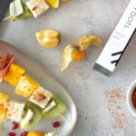 Obstspieße aus Melone, Mango, Ananas, Kiwi, Feta und Parmaschinken auf einem Teller. Nebendran liegt das Chilisalz von Roots mit dem die Obstspieße gewürzt sind