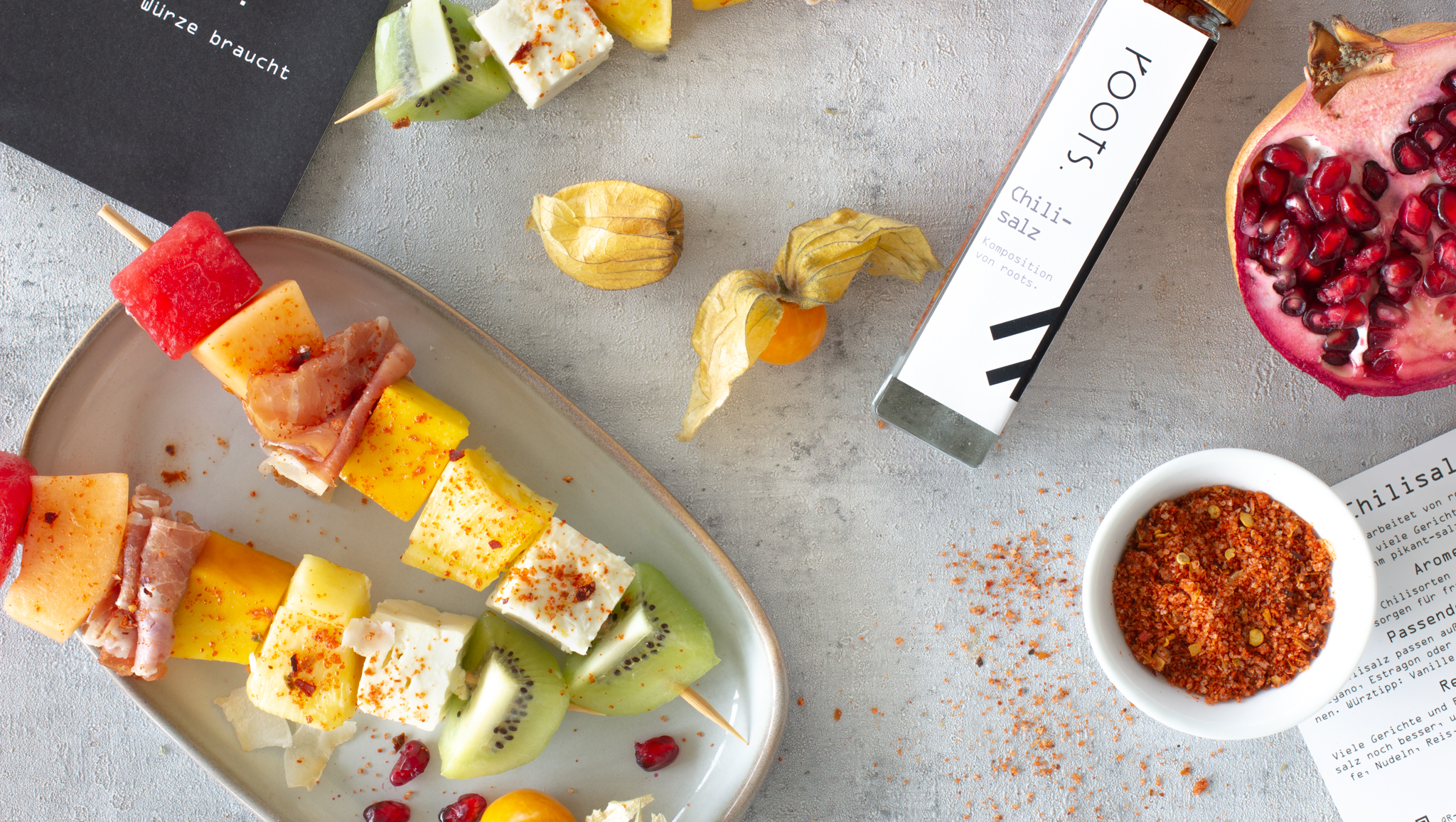 Obstspieße aus Melone, Mango, Ananas, Kiwi, Feta und Parmaschinken auf einem Teller. Nebendran liegt das Chilisalz von Roots mit dem die Obstspieße gewürzt sind
