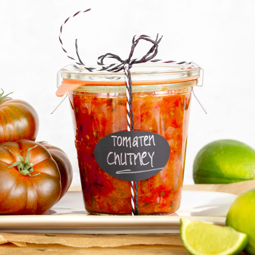 Ein Glas selbstgemachtes Tomaten-Chutney als Geschenk verpackt auf einem Teller, nebenan liegen Tomaten und Limetten