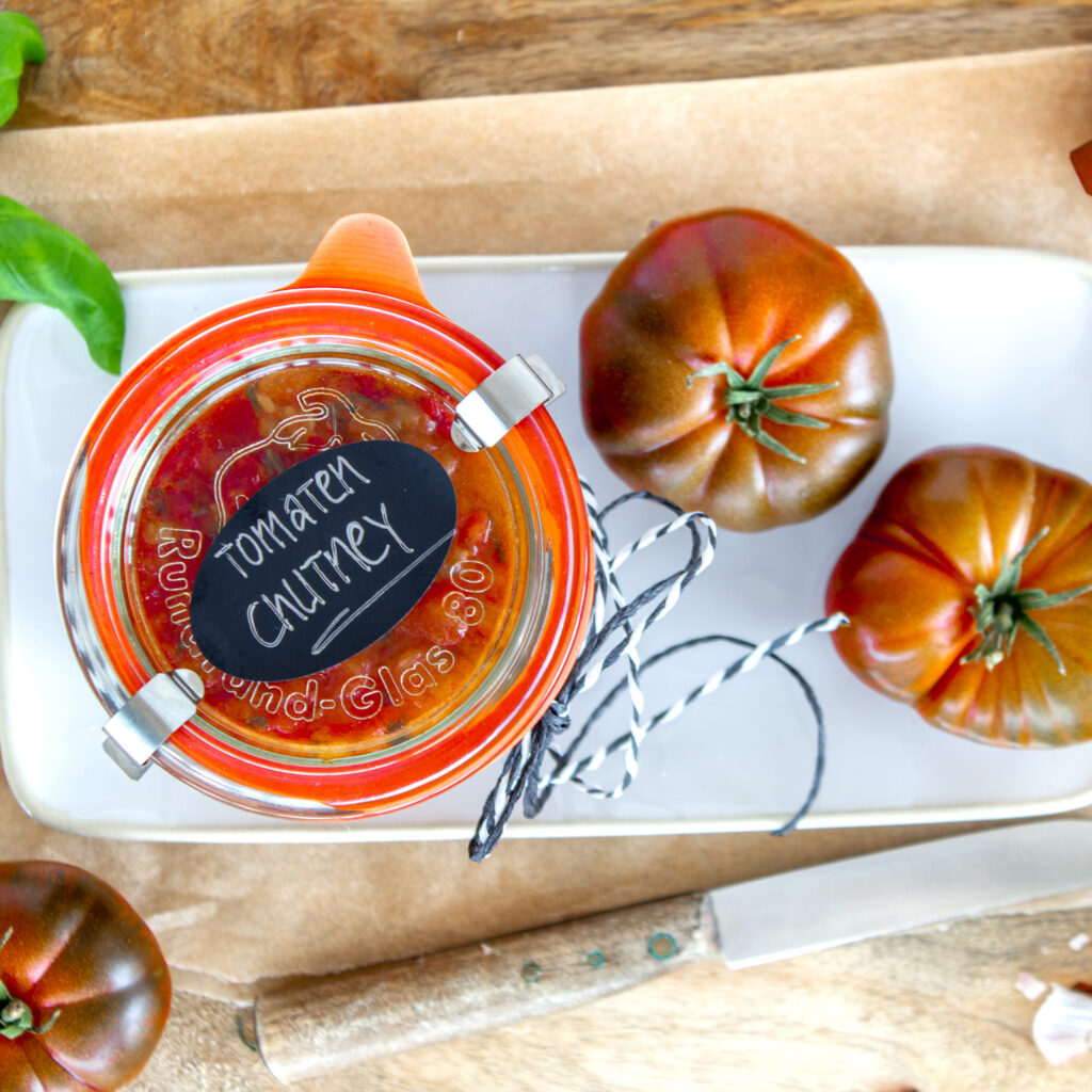 Ein Glas selbstgemachtes Tomaten-Chutney als Geschenk verpackt auf einem Teller, nebenan liegen zwei Tomaten