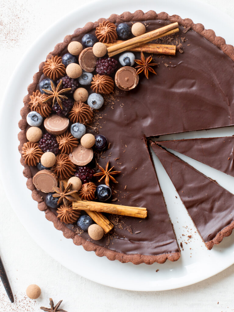Angeschnittene Schokoladen-Lebkuchen-Tarte mit opulentem edlem Topping aus Heidelbeeren, Brombeeren, Schokokugeln, Sternanis, Zimtstangen und Schokosplittern, ein zugeschnittenes Stück liegt nebenan