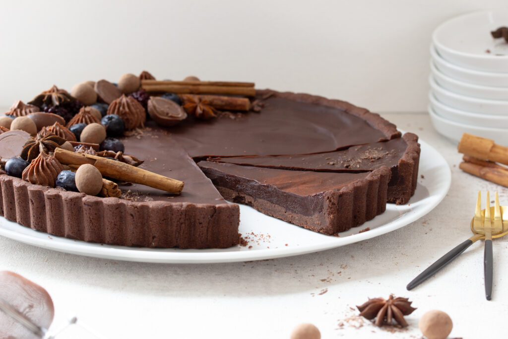 Angeschnittene Schokoladen-Lebkuchen-Tarte mit opulentem edlem Topping aus Heidelbeeren, Brombeeren, Schokokugeln, Sternanis, Zimtstangen und Schokosplittern, seitliche Sicht