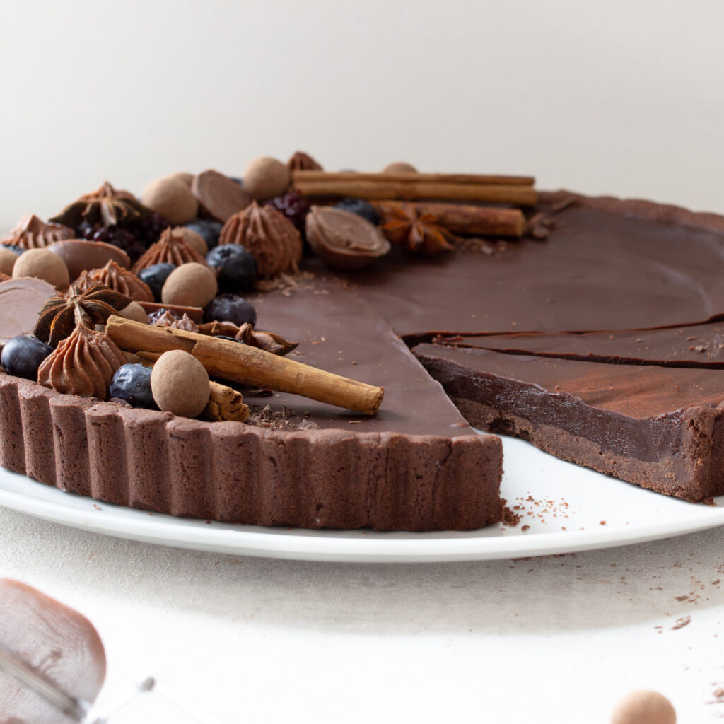 Angeschnittene Schokoladen-Lebkuchen-Tarte mit opulentem edlem Topping aus Heidelbeeren, Brombeeren, Schokokugeln, Sternanis, Zimtstangen und Schokosplittern, seitliche Sicht