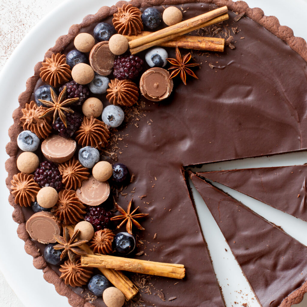 Angeschnittene Schokoladen-Lebkuchen-Tarte mit opulentem edlem Topping aus Heidelbeeren, Brombeeren, Schokokugeln, Sternanis, Zimtstangen und Schokosplittern, Vogelperspektive
